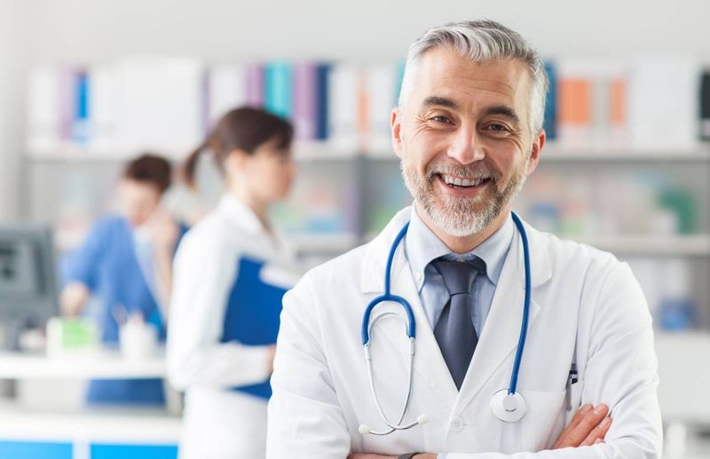 Smiling doctor posing with arms crossed in the office, he is wearing a stethoscope, medical staff on the background